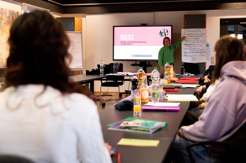 Lehrerin vor einer digitalen Leinwand und einer Flipchart; Schülerinnen und Schüler an Tischen, vor ihnen liegen Hefte