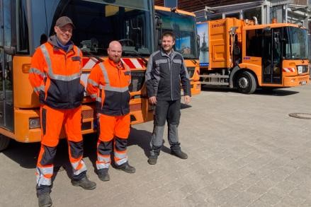 Drei junge Männer vor einem Müllfahrzeug, alle machen die „Daumen hoch“ Geste. Zwei der Männer tragen orangefarbene Warnschutzkleidung, der dritte Mann trägt schwarz-graue Arbeitskleidung eines Mechanikers.