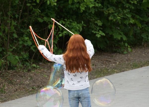 Zu sehen ist ein Mädchen von hinten mit langen Haaren, das große Seifenblasen mit einer Schnur macht