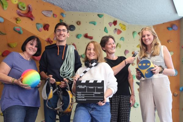 Fünf Personen stehen vor einer Kletterwand und halten verschieden Gegenstädne zu den Workshops in der Hand: Kabel, eine Kamerklappe und ein Mikrofon