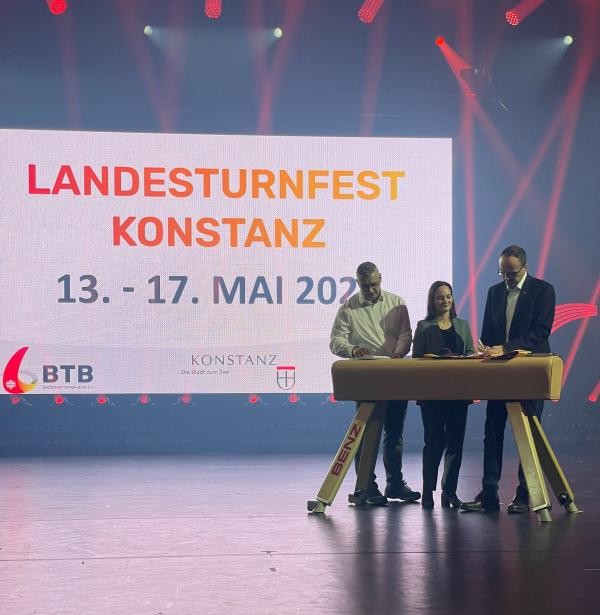 Drei personen stehen an eine Turn-Pferd und schreiben. Im HIntergrund sieht man eine Projektion mit Text: "Landesturnfest Konstanz 13. - 17. Mai 2026"