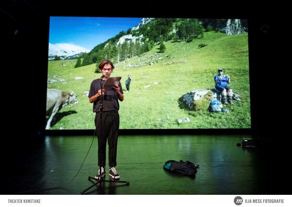 Eine Person steht vor einer Leinwand, auf der eine grüne Wiese zu sehen ist. Die Person steht an einem Mirko und liest etwas vor.