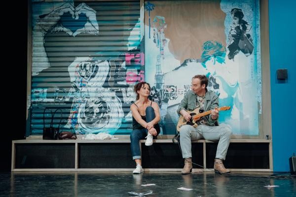 Eine Frau und ein Mann sitzen auf einer Bank, der Mann spielt Gitarre.