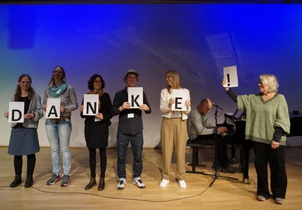 Eine Gruppe Personen, die je einen Buchstben des Wortes "Danke" halten. Im Hintergrund sitzt jemand an einem Klavier.