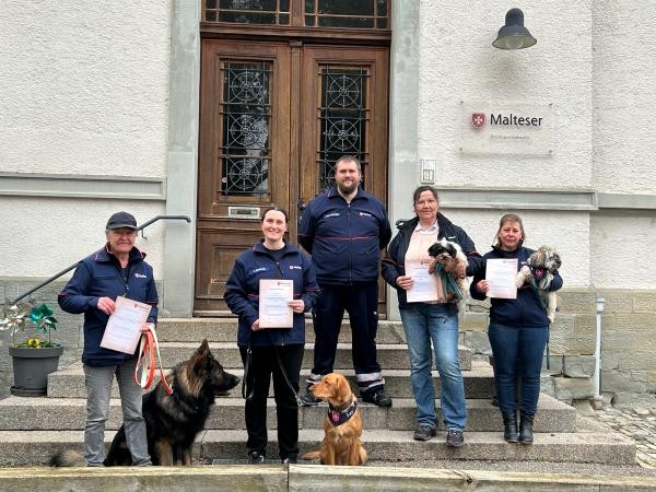 Mehrere Personen mit Hunden stehen auf einer Treppe vor dem Gebäude der Malteser und halten Dokumente in den Händen