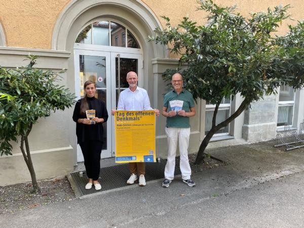 Drei Personen stehen vor einem Hauseingang und halten ein Plakat und Flyer in den Händen