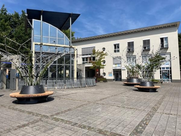 Pflanzkübel mit schirmartigen, begrünten Rankgittern und Sitzbänken drum herum stehen auf einem Platz mit steinernen Bodenplatten