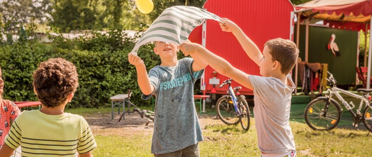 Kinder spielen mit Tuch und Ball auf einer Wiese vor einem bunten Bauwagen